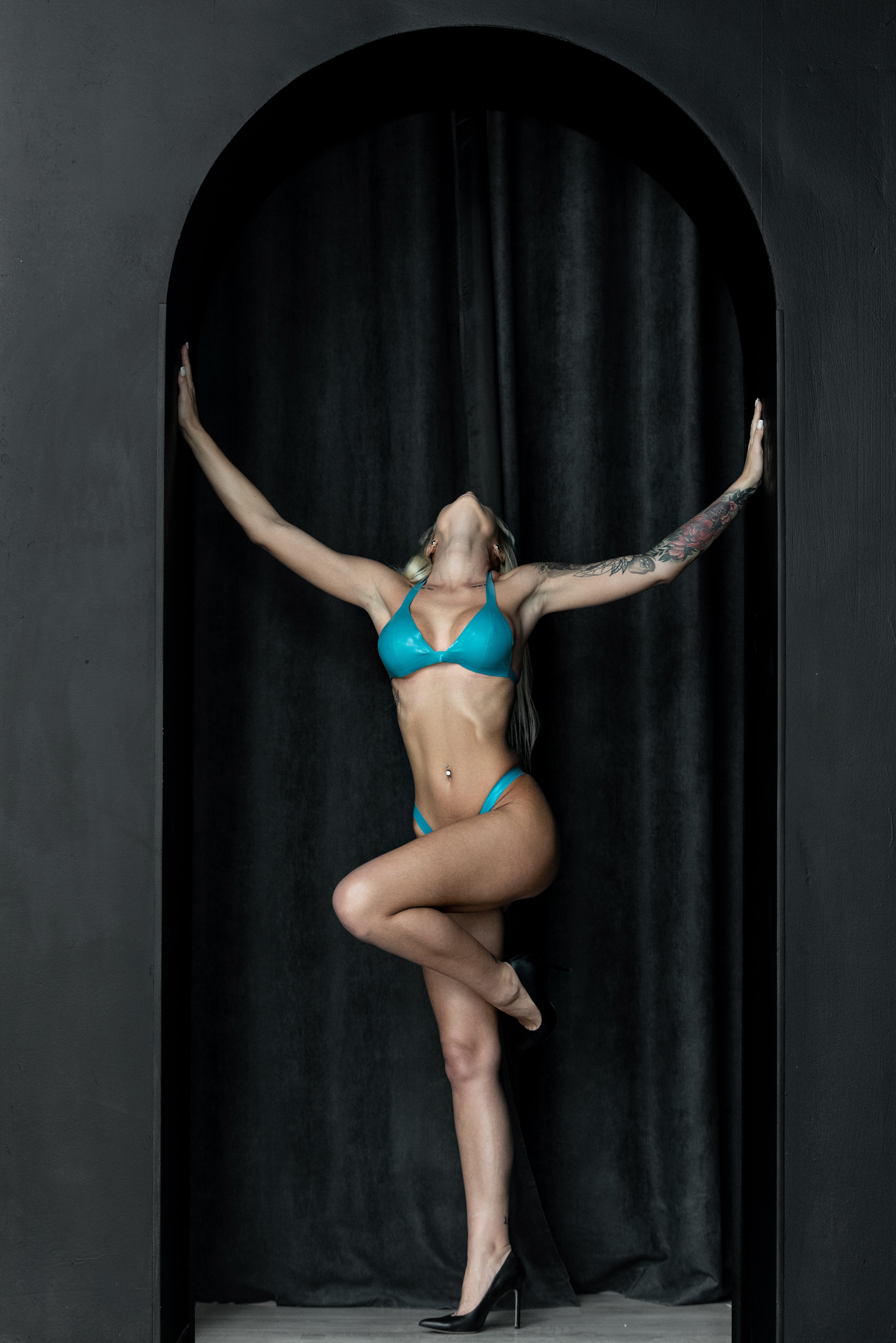 Person wearing a blue latex bra and panties standing with arms outstretched against a black curtain.