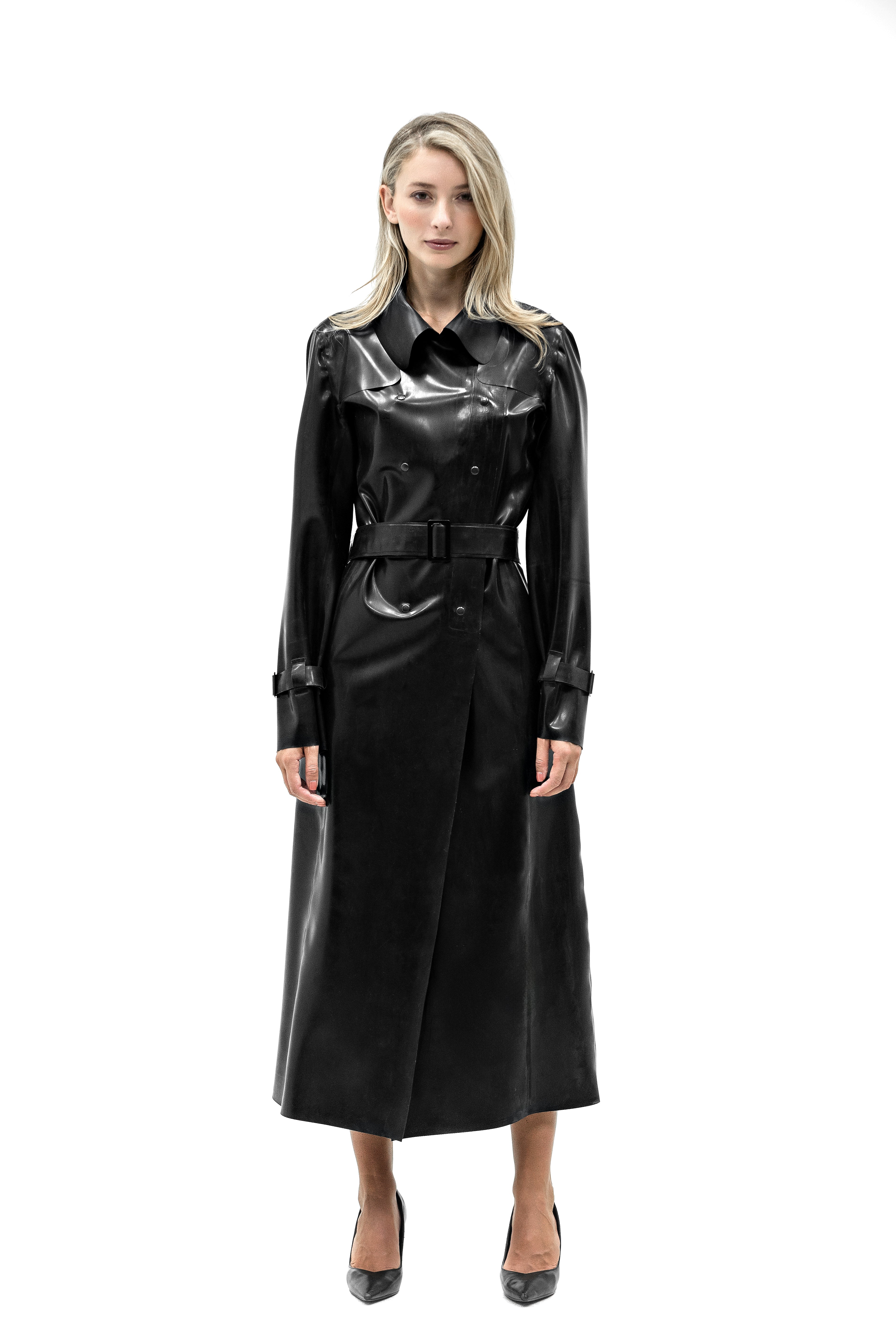 A woman modeling a long Latex black trench coat with a glossy, patent-like finish on the chest and sleeves, paired with a matching belt and black pointed-toe heels, shot against a solid white background.
