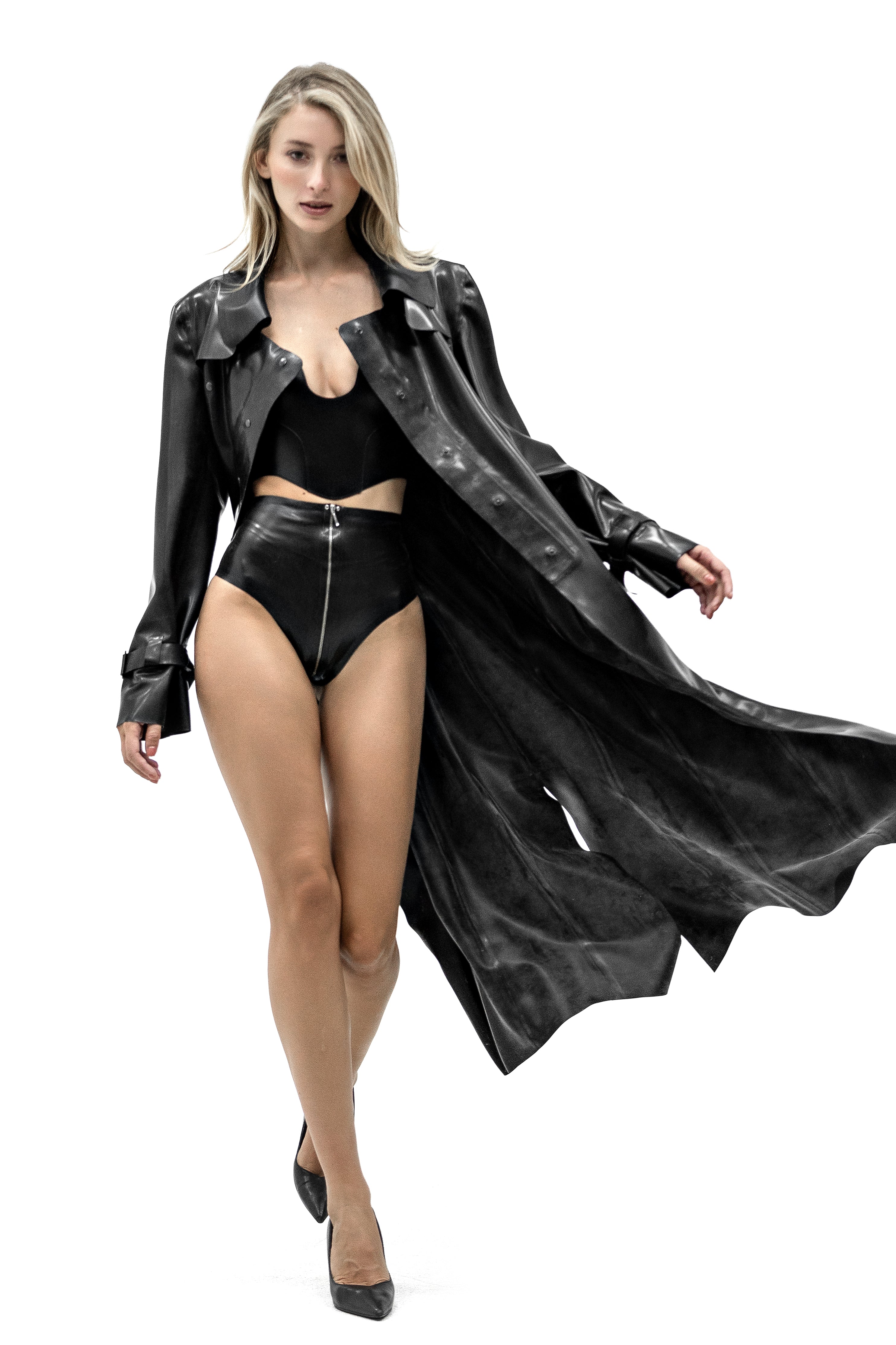 A woman modeling a black Latex faux-leather trench coat over a black two-piece swimsuit and black heels, captured against a solid white background.